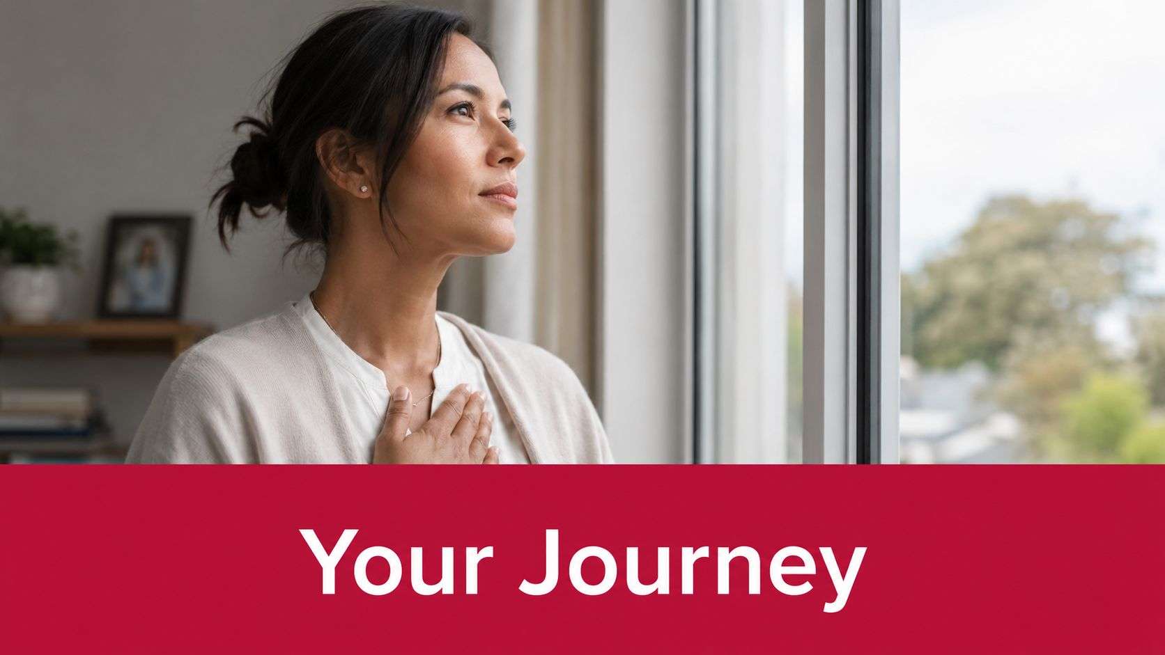 A woman looks reflectively out a window with her hand placed gently over her heart.